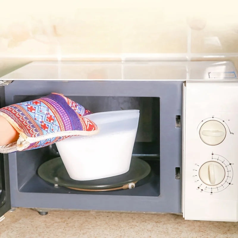 Hand with colorful oven mitt removing a white reusable silicone food storage bag from a microwave after heating contents