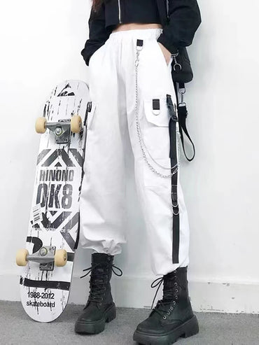 Woman wearing white Harajuku-style cargo joggers with chains, black combat boots, and a skateboard, showcasing edgy women's streetwear.