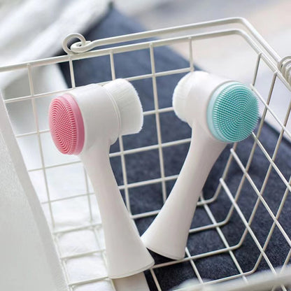 Two dual-sided silicone face brushes with soft bristles and textured massage heads (pink and blue), placed inside a white wire basket on a towel