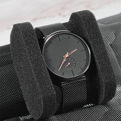 Close-up of black mesh wristwatch with rose gold hands on a soft black foam insert, displayed on a textured watch case
