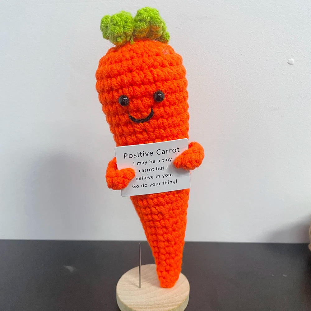 Handmade crochet carrot plush doll with smiling face, holding a sign that says "Positive Carrot – I may be a tiny carrot, but I believe in you. Go do your thing!