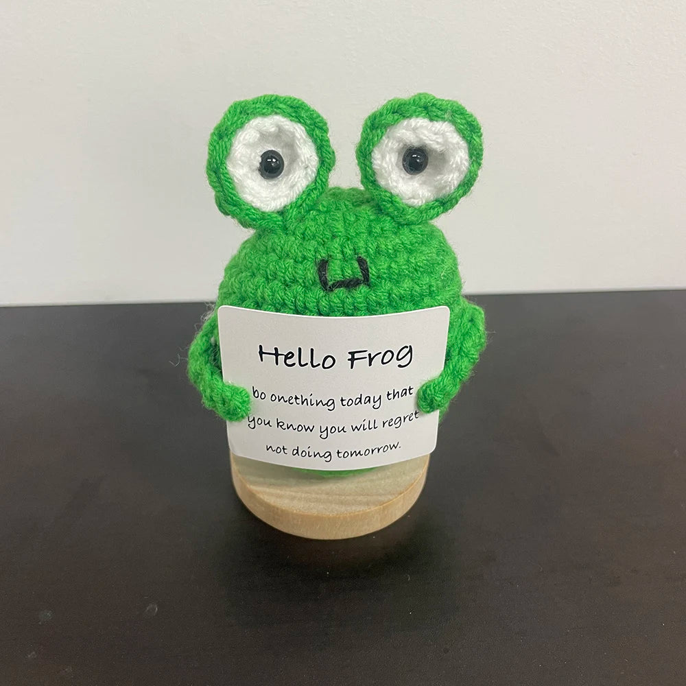 Handmade crochet frog plush with large eyes and a smiling face, holding a sign that says "Hello Frog – Do something today that you know you will regret not doing tomorrow.