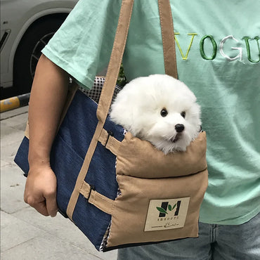 Small white dog sitting in a portable car seat bag carried by its owner outdoors.
