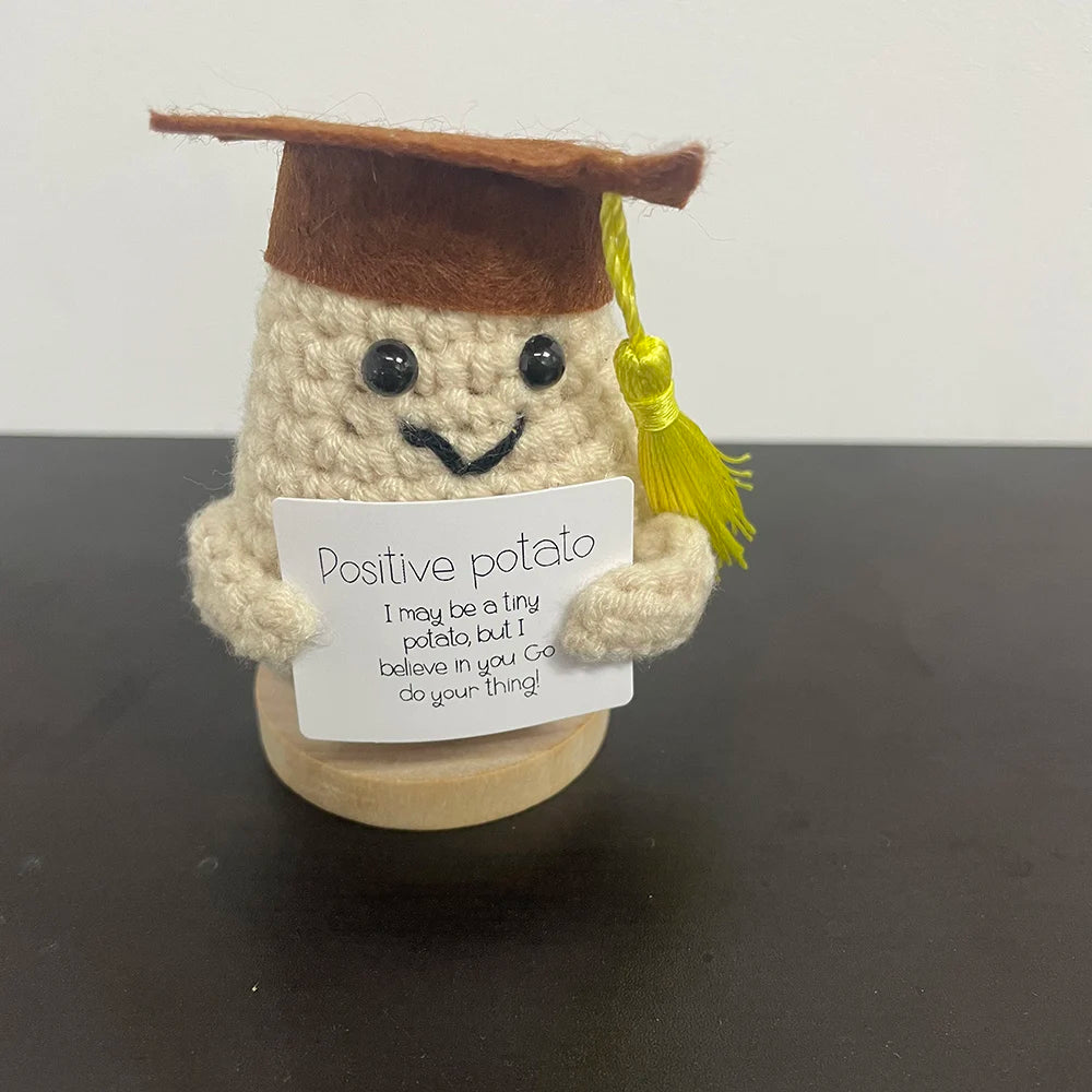 Handmade crochet potato plush doll wearing a graduation cap with a yellow tassel, holding a sign that says "Positive potato – I may be a tiny potato, but I believe in you. Go do your thing!