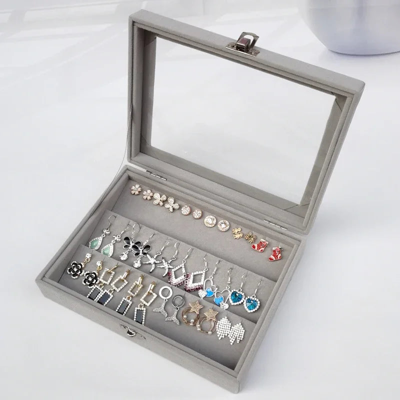 Gray velvet jewelry display box with transparent lid showing assorted colorful earrings in various designs
