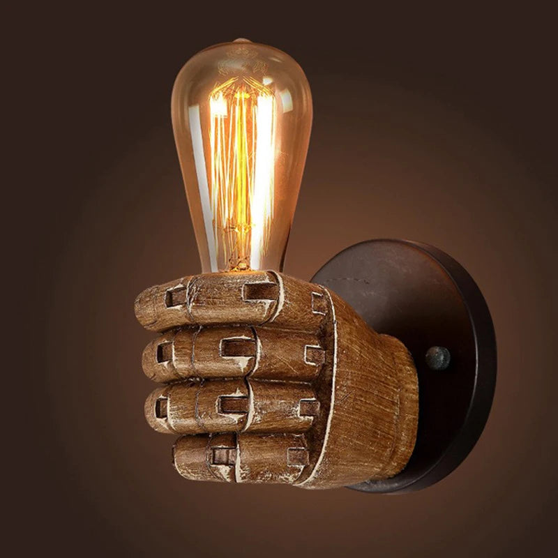 Industrial-style resin wall lamp shaped like a wooden hand gripping a vertical Edison bulb, mounted on a round black base with warm ambient lighting