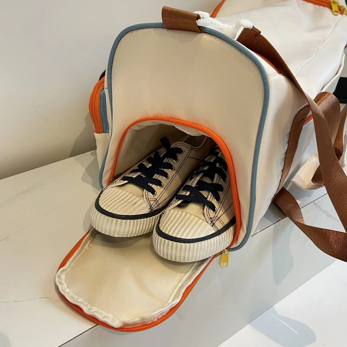White nylon duffel bag with open side shoe compartment holding sneakers, featuring orange zipper and brown shoulder straps