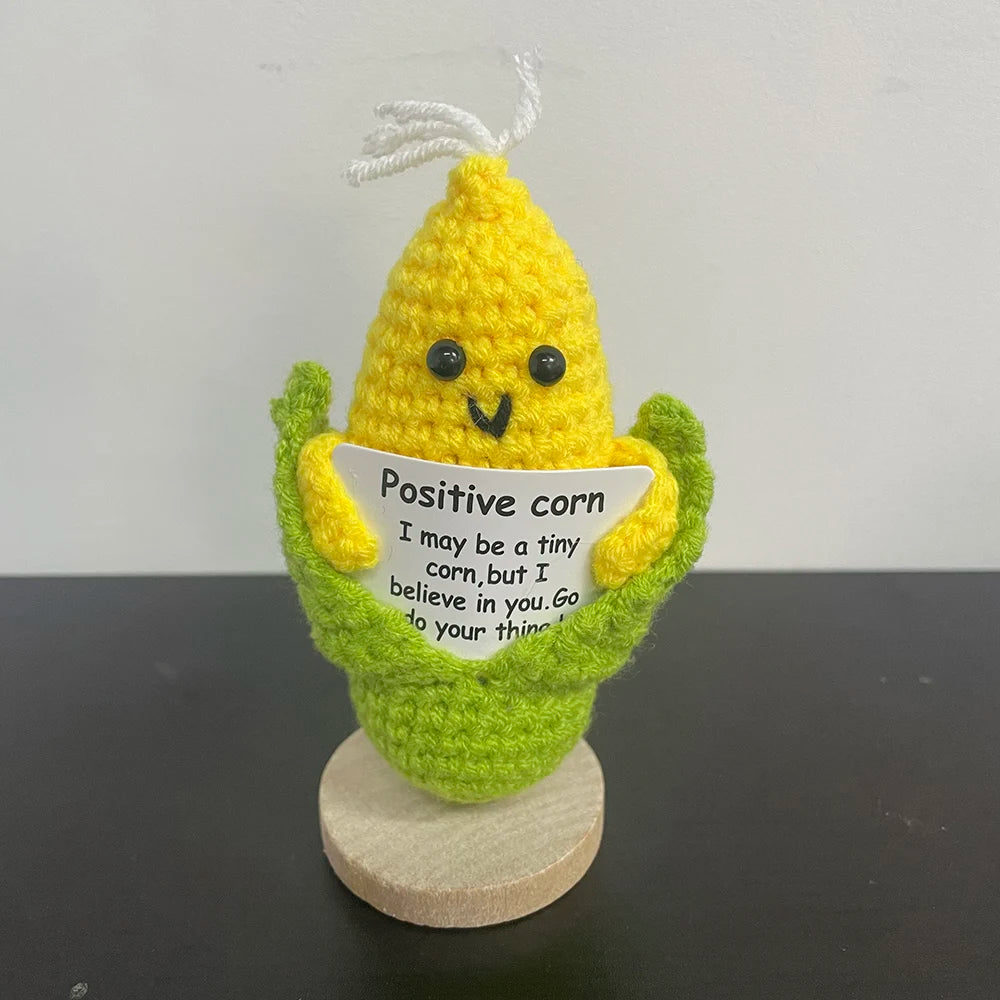Handmade crochet corn plush doll with husks and a smiling face, holding a sign that says "Positive corn – I may be a tiny corn, but I believe in you. Go do your thing!