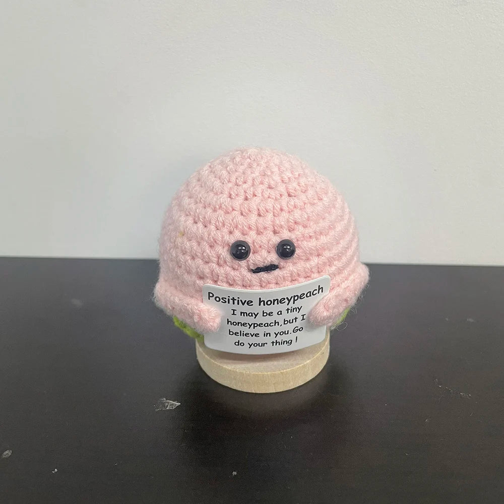 Handmade crochet honeypeach plush doll in soft pink yarn, holding a sign that says "Positive honeypeach – I may be a tiny honeypeach, but I believe in you. Go do your thing!