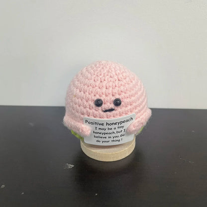 Handmade crochet honeypeach plush doll in soft pink yarn, holding a sign that says "Positive honeypeach – I may be a tiny honeypeach, but I believe in you. Go do your thing!