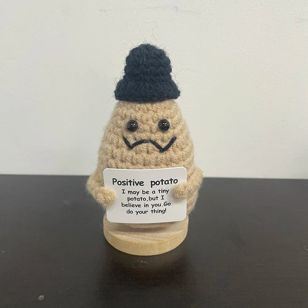 Handmade crochet potato plush doll wearing a black beanie with a frowning expression, holding a sign that says "Positive potato – I may be a tiny potato, but I believe in you. Go do your thing!