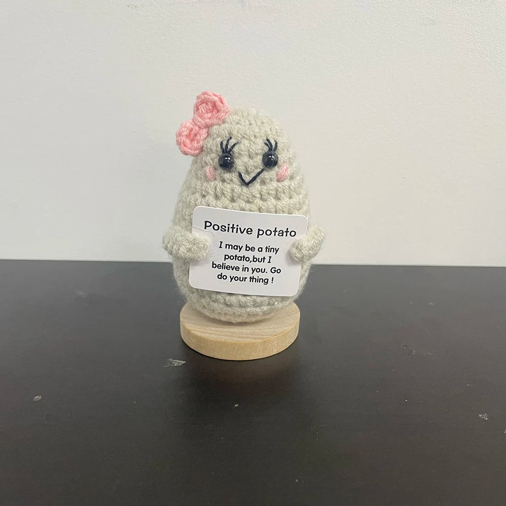 Handmade light gray crochet potato plush with pink bow and eyelashes, holding a sign that says "Positive potato – I may be a tiny potato, but I believe in you. Go do your thing!