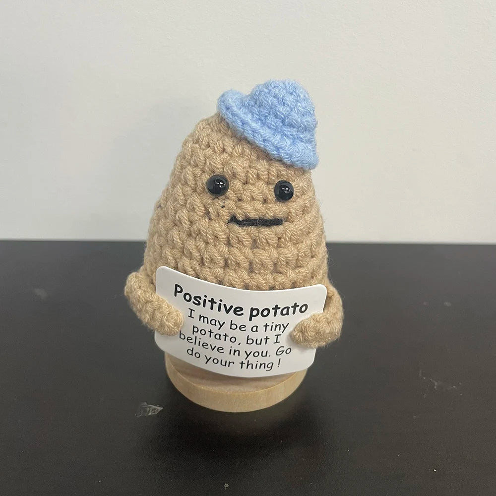 Handmade crochet plush of a light brown potato wearing a light blue yarn hat, holding a motivational sign that reads "Positive potato – I may be a tiny potato, but I believe in you. Go do your thing!
