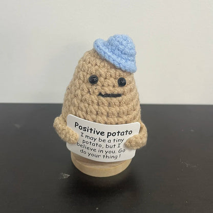 Handmade crochet plush of a light brown potato wearing a light blue yarn hat, holding a motivational sign that reads "Positive potato – I may be a tiny potato, but I believe in you. Go do your thing!