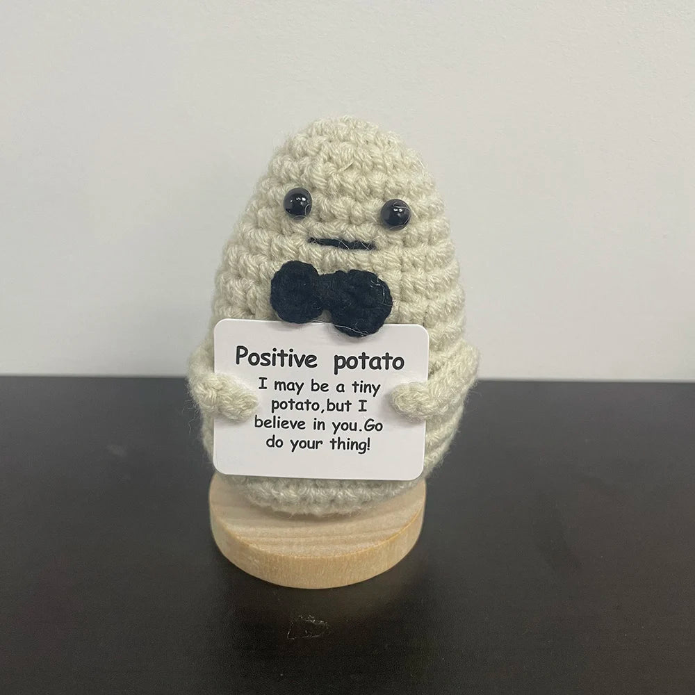 Handmade crochet potato plush doll in light gray yarn wearing a black felt bow tie, holding a sign that says "Positive potato – I may be a tiny potato, but I believe in you. Go do your thing!