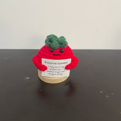 Red crochet tomato plush doll with green top, holding a sign that says "Positive tomato – I may be a tiny tomato, but I believe in you. Go do your thing!