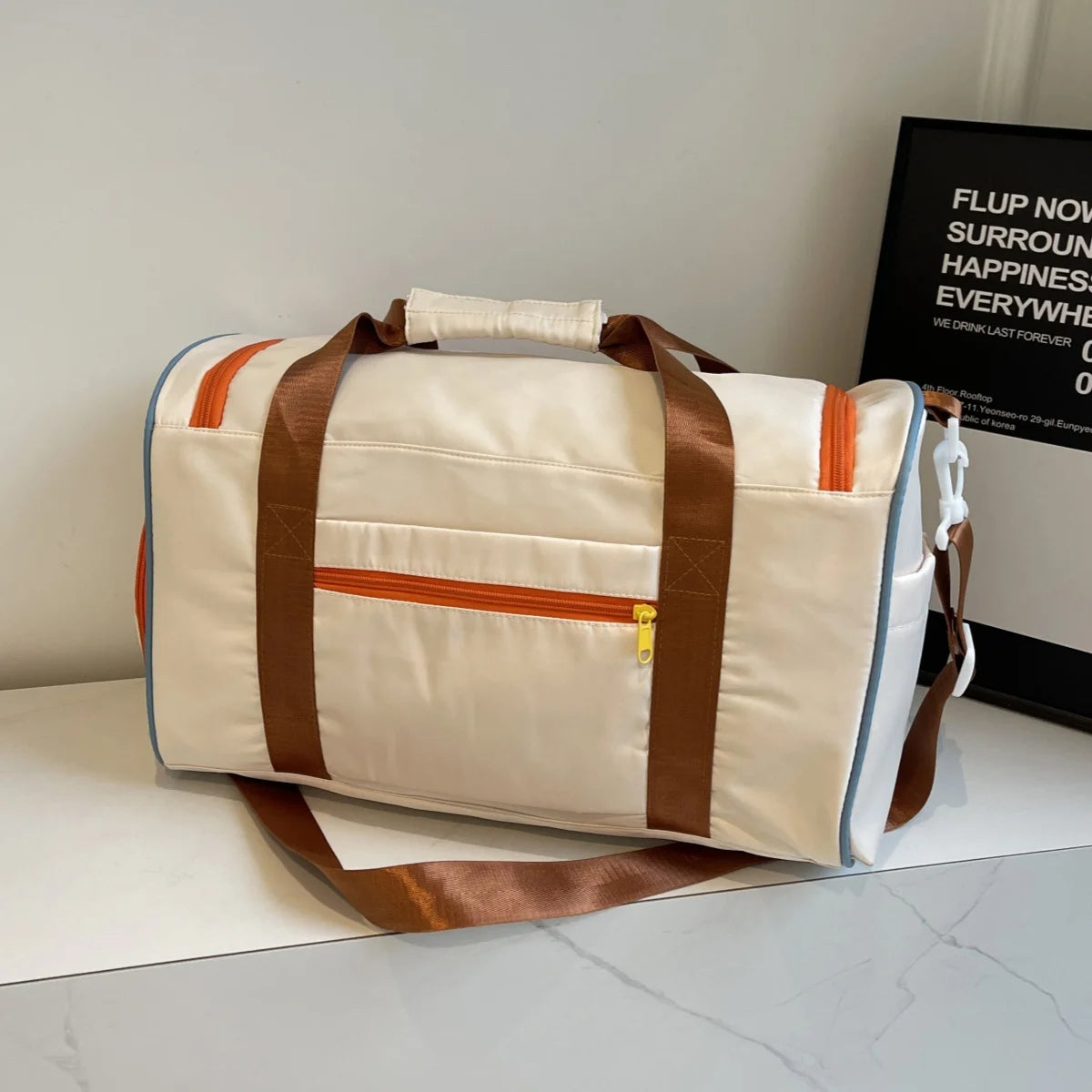 Back view of white nylon duffel bag with brown shoulder straps and orange zipper pocket, placed on a table near framed decor
