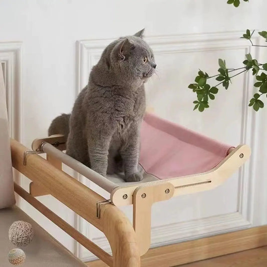 Gray cat sitting on a pink canvas-lined window-mounted hammock bed with a wooden frame, designed for comfort and elevated views.